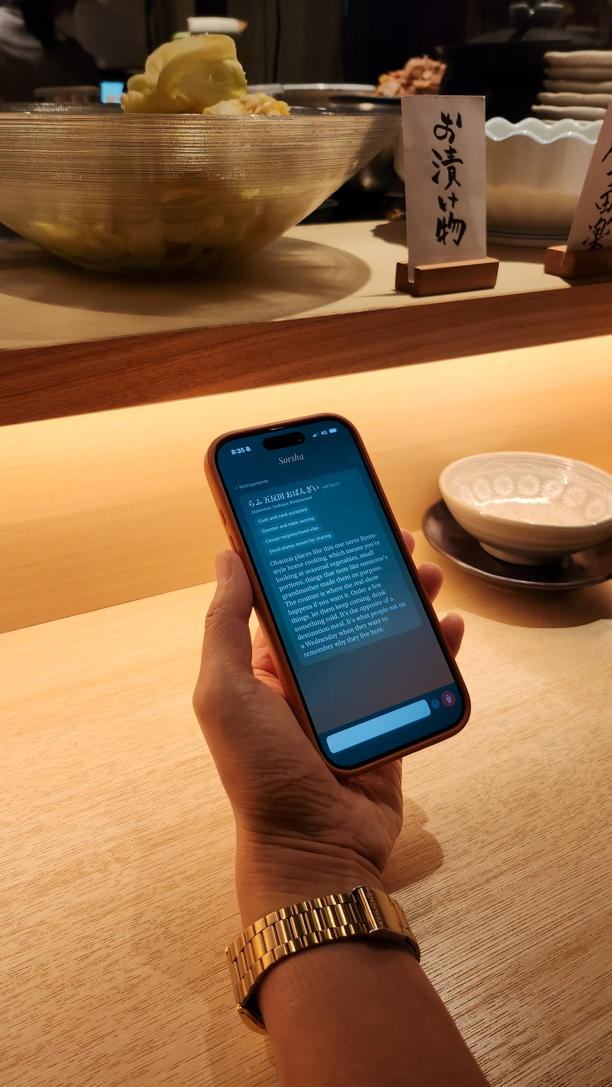 A hand holding a phone at a restaurant counter in Tokyo, with the Sorsha brief on screen